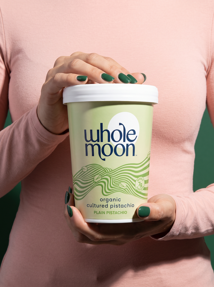 A person in a pink long-sleeve top holds a tub of Whole Moon organic cultured pistachio yogurt. The container is light green with dark blue branding and green wave graphics, topped with a white lid.