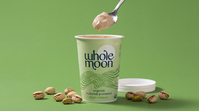 A Whole Moon organic cultured pistachio yogurt cup on a green background, with a spoon lifting some of the yogurt above the cup and pistachios scattered around it.