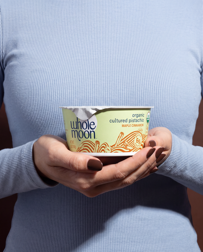 A person in a light blue ribbed sweater holds a cup of Whole Moon Organic Cultured Pistachio Maple Cinnamon yogurt against a dark brown background.