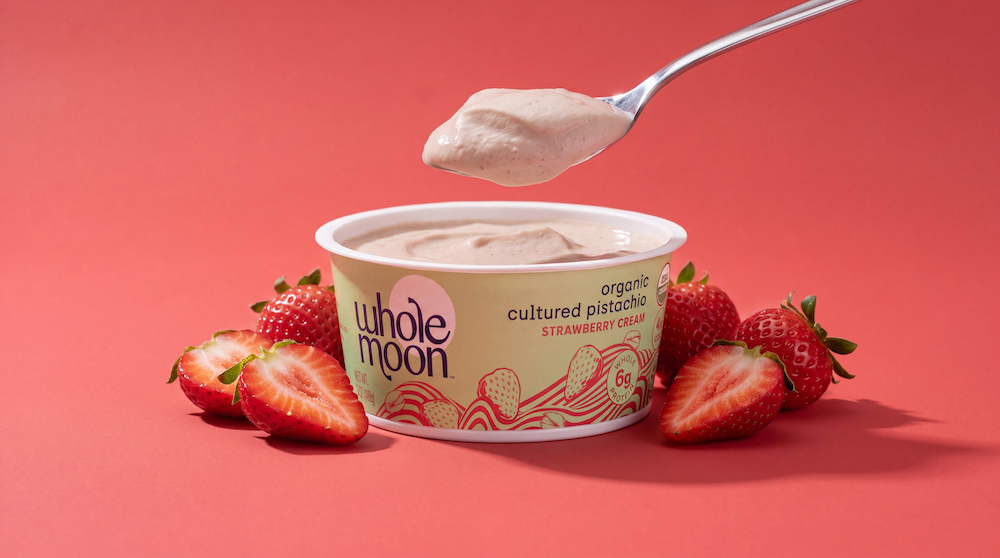 A tub of Whole Moon Organic Cultured Pistachio Strawberry yogurt sits on a coral-red background, surrounded by fresh strawberries and a spoon holding a scoop of the creamy product above the container.
