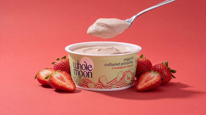 A tub of Whole Moon Organic Cultured Pistachio Strawberry yogurt sits on a coral-red background, surrounded by fresh strawberries and a spoon holding a scoop of the creamy product above the container.