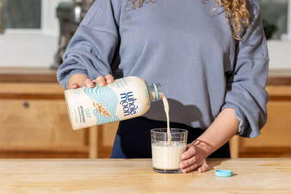 Woman pouring Wholemoon whole protein oat milk from beige carton into a glass, creating a smooth white stream against a bright kitchen background