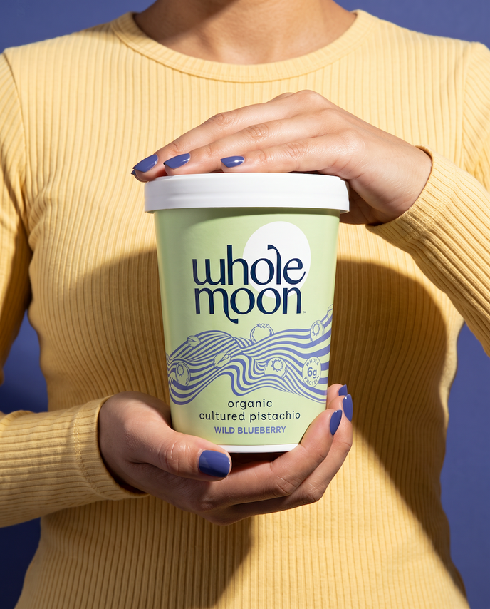 A person in a ribbed yellow sweater holds a Whole Moon organic cultured pistachio Wild Blueberry ice cream tub. The tub is light green with blue wave graphics and a white lid, set against a solid blue background.