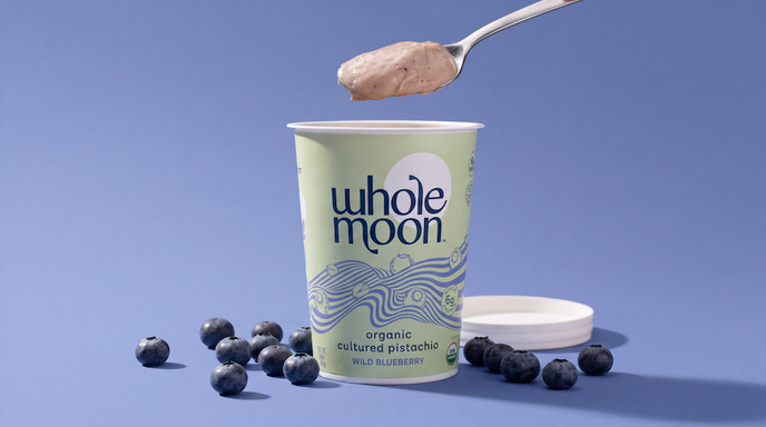 A Whole Moon organic cultured pistachio yogurt cup on a blue background, with a spoon above holding a scoop of yogurt and blueberries scattered around the cup.