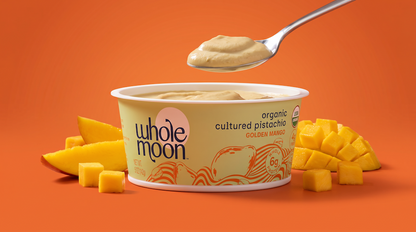 A Whole Moon organic cultured pistachio yogurt tub in Golden Mango flavor, shown on a bright orange background with a spoonful of the creamy yellow-green yogurt above and mango slices and cubes around the container.