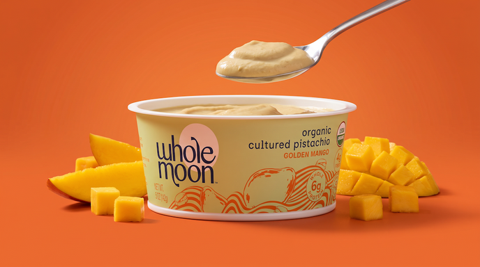 A Whole Moon organic cultured pistachio yogurt tub in Golden Mango flavor, shown on a bright orange background with a spoonful of the creamy yellow-green yogurt above and mango slices and cubes around the container.