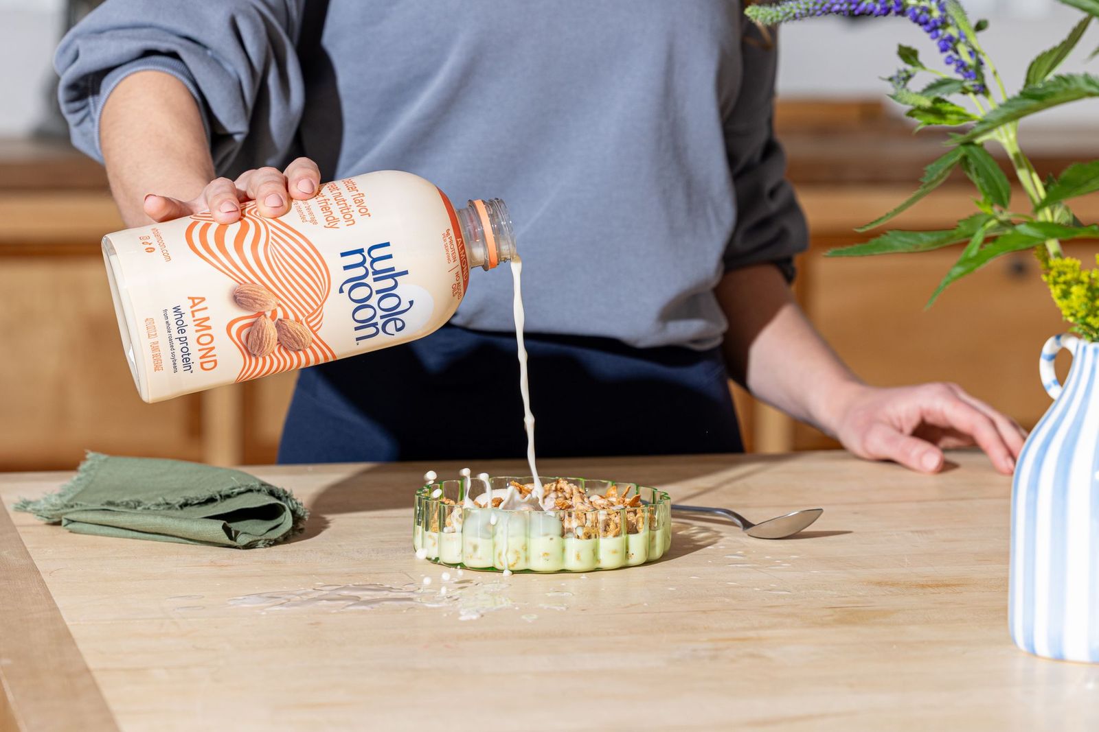 Woman pouring Wholemoon whole protein almond milk from beige carton into a cereal bowl, creating a smooth white stream against a bright kitchen background