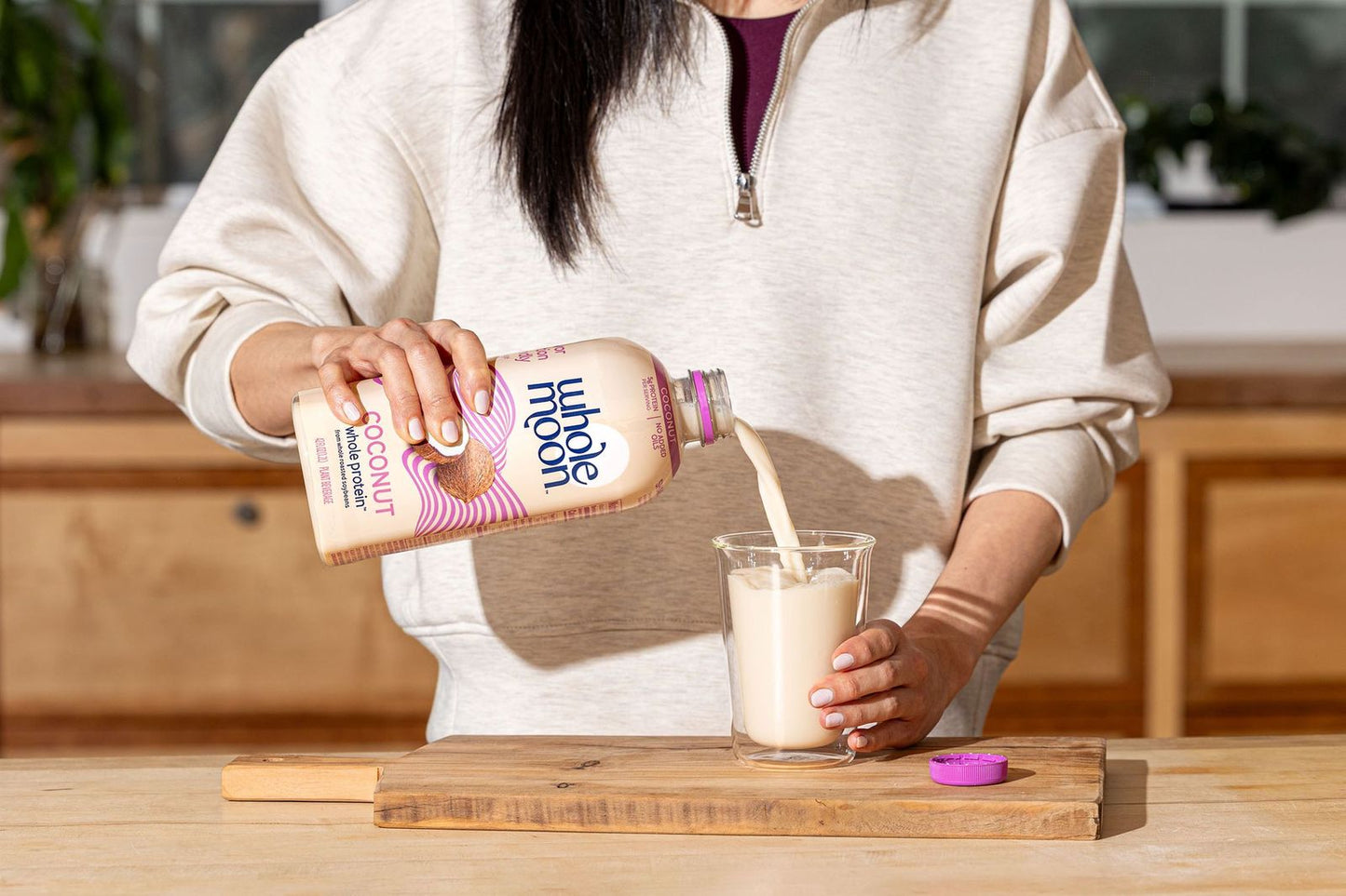 Woman pouring Wholemoon whole protein coconut milk from beige carton into a glass, creating a smooth white stream against a bright kitchen background