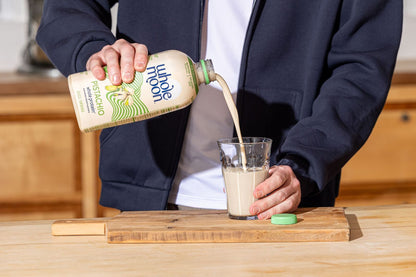Man pouring Wholemoon whole protein pistachio milk from beige carton into a glass, creating a smooth white stream against a bright kitchen background